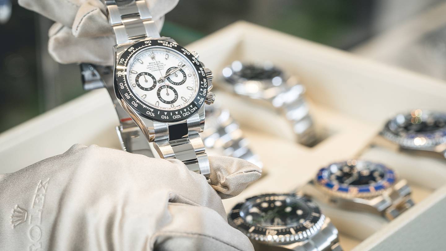 A Rolex watch being held in white gloves to show a customer, above a box full of silver watches.