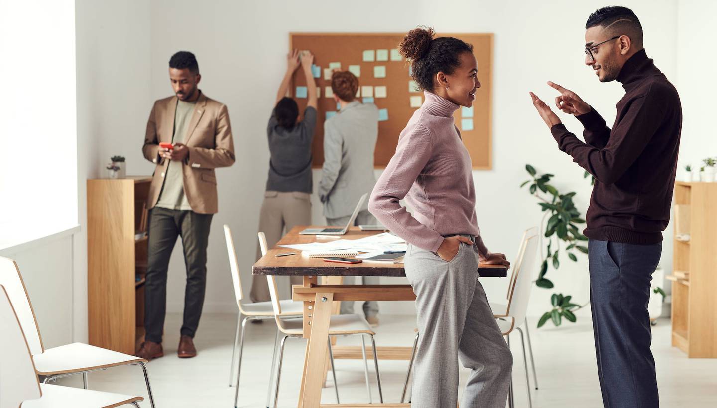 Fashion professionals stand to discuss a topic while colleagues in the background work before a pinboard.